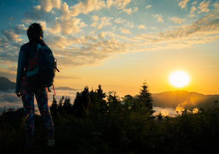 Young fit happy woman stand on viewpoint after reach top in mountains. Carefree tourist woman looking at sun enjoying landscape. Girl traveler on top of mountain in rays of sunsetの写真素材