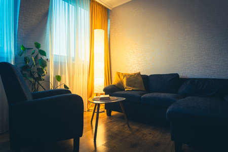 Cozy apartment living room with sofa and yellow pillows and stylish table with book by flower and window with blue light outside in winter. Early evening book read. Copy paste wallの写真素材