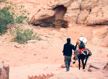 Petra, Jordan - 7th october, 2022: bedouin master with donkey take overweight Petra female visitor client down stairs from little Petra - monastery uphillのeditorial素材