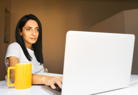 Young caucasian woman sits in apartment behind white desktop and look straight ahead. Concept of work from home.の写真素材