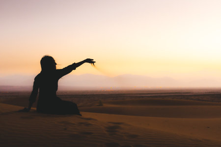 Traveler explorer girl sit in Desert and let sand fall from the hand.Silhouette of woman playing with sand on sunset in summer vacation as twilight. Hand pours sandの写真素材