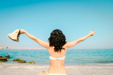 Vacation and freedom. Happy young brunette woman rising hands up enjoy standing on tropical beach enjoying beautiful view hold summer hat from behind. Carefree lifestyle conceptの写真素材