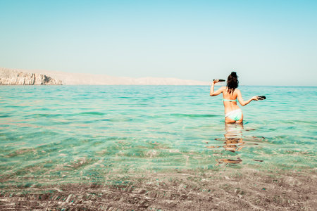 Young carefree Caucasian woman tourist dance on the beach in turquoise water alone with hands up hold slippers on summer vacation beach holidays. Solo travel adventure wanderlustの写真素材