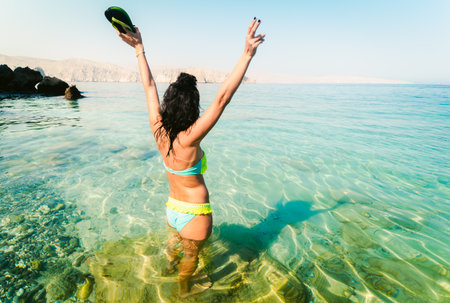Young carefree caucasian woman stand excited in turquoise water beach alone with hands up hold slippers on summer vacation beach holidays.Solo holidays vacation in middle eastの写真素材