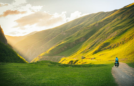 Cyclist back view on the road in green caucasus nature with scenic sunset background on valley. Traveller on bicycle. Solo travel long distance bicycle touring. Off-road bikingの写真素材