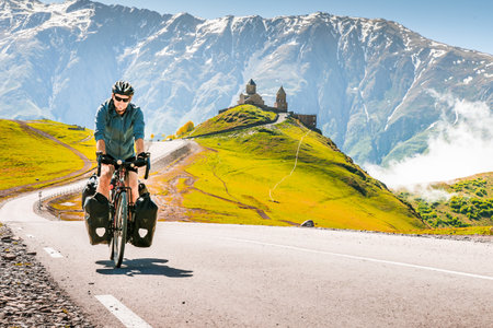 Cyclist back view on the road in scenic caucasus nature with Gergeti trinity monastery in the background. Traveller on bicycle. Solo travel long distance bicycle touring conceptの写真素材