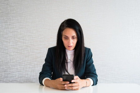 Young caucasian brunette business woman director in office hold smartphone read news surprised in shock at workの写真素材