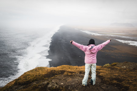 Young woman in pink jacket stand on top of mountain summit raises arms into air, happy and drunk on...