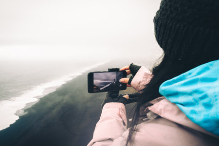 Front view of caucasian beautiful woman wearing winter clothes taking pictures selfie with black sand beach in in Icelandの写真素材