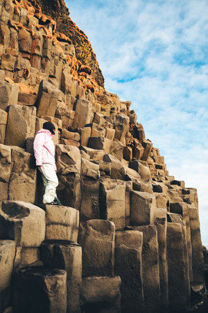 Adult caucasian woman tourist in yellow jacket standing on basalt columns on Reynisfjara Black Sand Beach in Icelandの写真素材