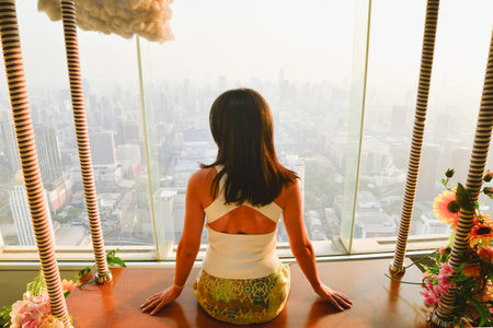 Pretty caucasian woman tourist stand behind glass window isolated enjoying beautiful sunset panorama. Sky view Thailandâs capital Bangkok cityscapeの写真素材