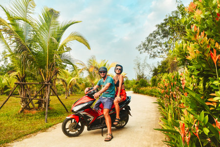 Happy smiling couple travelers riding motorbike scooter in safety helmets during tropical vacation under palm trees on Phu Quoc island , Vietnamの写真素材