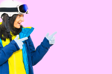 Skier young woman wearing warm padded windbreaker jacket hat ski goggles mask point index finger aside on copy space blank area travel rest spend weekend winter season. Isolated on pink backgroundの写真素材