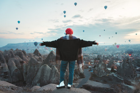 Close up aerial zoom out view panorama caucasian Woman solo traveler on viewpoint rising her hands and looking on flight hot air balloons in Cappadocia. Love valley sunrise in cold winter morningの写真素材