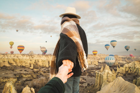 Couple hold hand show looking admire hot air balloon show ,wear fashionable clothing stand on scenic viewpoint watch hot air balloon flight on sunrise with beautiful goreme town and valley backgroundの写真素材