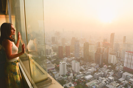 Pretty caucasian woman tourist stand behind glass window isolated enjoying beautiful sunset panorama. Sky view Thailandâs capital Bangkok cityscapeの写真素材