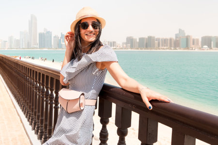 Portrait caucasian pretty tourist woman at viewpoint looking at camera pose with Abu Dhabi skyline panorama and beach, reflecting peace, relaxation, and urban beauty in the UAEの写真素材