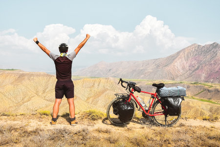 Adventurous muscular young arms outstretched caucasian young male cyclist on viewpoint by red touring bicycle in deserted mountains with hands up confident carefree lifestyleの写真素材