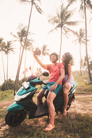 Happy cheerful young caucasian couple sit on scooter enjoy sunset taking selfie posting on social media Sri Lanka. Embracing freedom and romance, road trip with tropical landscapes background at dawnの写真素材