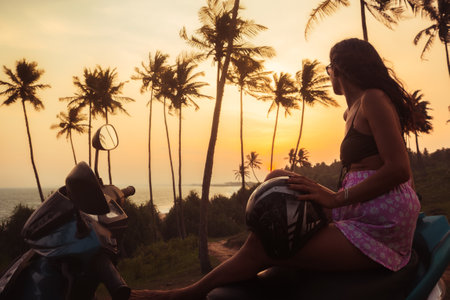 Carefree pretty woman on sit scooter by coastal viewpoint watch indian ocean and setting sun on sunset in Sri Lanka. Embracing freedom and romance while roaming tropical landscapes bathed in golden lightの写真素材