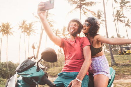 Carefree caucasian couple sit on scooter enjoy sunset taking selfie posting on social media Sri Lanka. Embracing freedom and romance, road trip with tropical landscapes background at dawnの写真素材