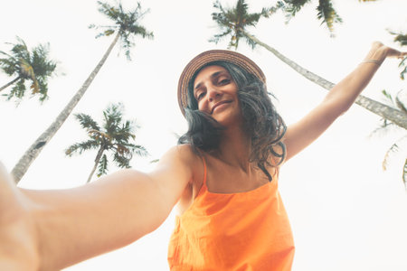 Casual Portrait beautiful Smiling happy vegan woman in orange dress straw hat takes selfie showing peace sign, framed by tall tropical palm trees bright sunny sky view.Slim body and healthy lifestyleの写真素材