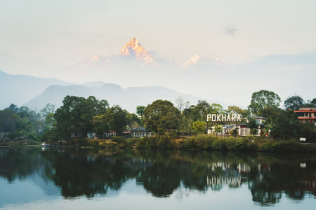 Scenic view of Pokhara city lakeside reflecting in calm water, with Machapuchare Fishtail peak glowing in morning light above misty Himalayan foothillsの写真素材