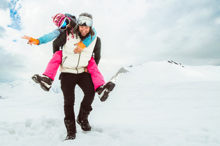 Joyful young couple walk piggyback in deep snow pose looking at camera in snowy mountain,...