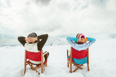 Back view couple sitting in deck chairs on snow hands behind heads enjoying mountain view winter leisureの写真素材