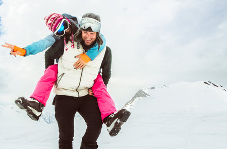 Joyful young couple walk piggyback in deep snow pose looking at camera in snowy mountain, symbolizing love, happiness, freedom, playful relationship, adventure, carefree winter escape, togethernessの写真素材