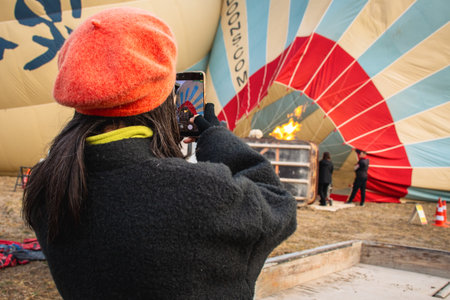 Back view woman in red beret capturing smartphone photo of colorful hot air balloon being inflated with burner flameの写真素材