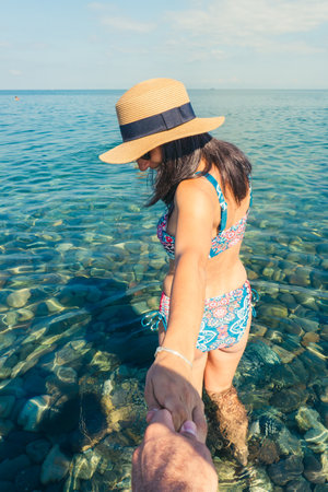 Young woman in summer hat and swimsuit holding partners hand in iconic follow me pose on crystal...