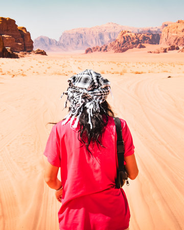 Back view of traveler wearing keffiyeh walking in Wadi Rum desert, Jordan, with red sand dunes and dramatic rock formations, famous UNESCO World Heritage Site and adventure travel destination.の写真素材