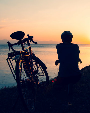 Traveler male cyclist rests beside bicycle at lake shore during golden sunset, concept of endurance,...
