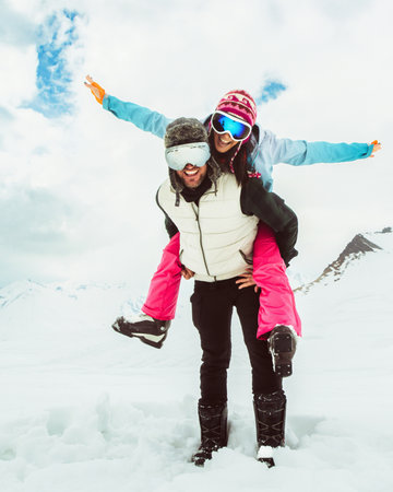 Joyful young couple walk piggyback in deep snow pose looking at camera in snowy mountain, symbolizing love, happiness, freedom, playful relationship, adventure, carefree winter escape, togethernessの写真素材