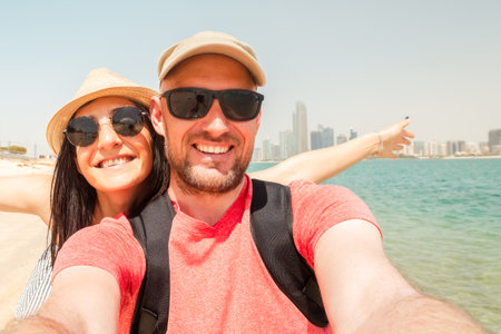 Couple of tourists having fun wave take selfie in Abu dhabi city beach on holidays in emirates - Happy friends laughing together on vacation - People and holidays concept. Explore sightseeing emiratesの写真素材