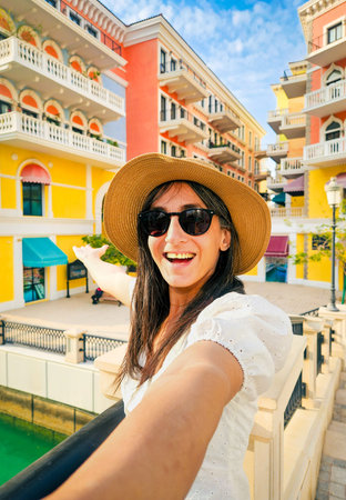 Excited Woman portrait smile taking smart phone selfie photo in Qanat Quartier Pearl Qatar colorful Venice style architecture with canals bridges luxury waterfront lifestyle travel destinationの写真素材