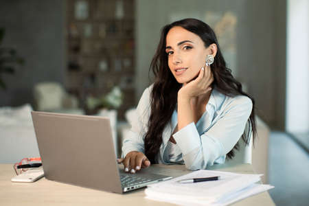 Smiling successful female using laptop computer indoors at home office, making notes on papers,...