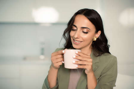 Smiling woman holding mug of coffee or tea, indoors at home in apartment, enjoying leisure time,...