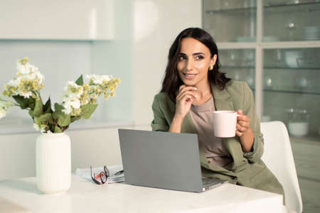 Business woman using laptop computer indoors at home office, with notes on papers, holding mug of...