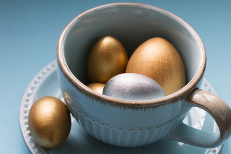 Gold  and silver Easter eggs in a porcelain mug on a blue background. National holiday celebration concepts. Happy Easter. Close up. Side view.の写真素材
