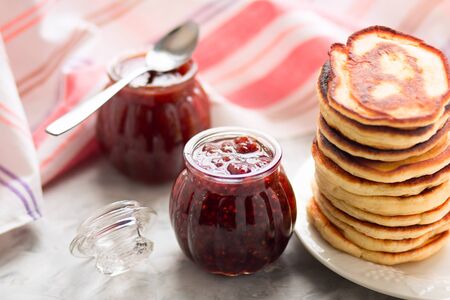Pancakes  on white plate and two jars of raspberry jam on the table. Selective focus. Front view