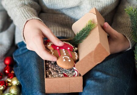 Girl hands pack Christmas gift, put in gift box gingerbread man, fir-tree branch, striped Lollipop...