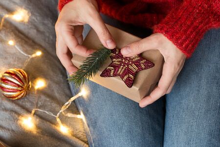 Female hands decorate gift box with red star and fir branch. Garland lights. Top view. Christmas,...