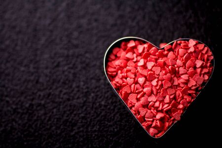 A metal cookie mold filled with small sweet candy hearts on a black surface. The concept of Valentines day. Top view. Macro. Copy spaceの写真素材