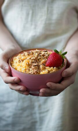 Pink plate of corn porridge with granola and strawberry in the hands of a girl. Healthy, diet, vegan or food concept for social media. Close up. Front viewの写真素材