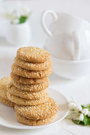 Freshly baked round sesame cookies with a apple tree flowers and tea cups on a white table. Cook at...