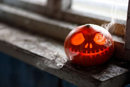 Smoking Halloween pumpkin on the dirty window sill of an old house window. Big spooky helloween symbol. Selective focusの写真素材