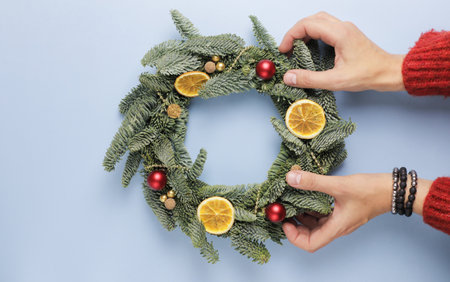 Womens hands hold over a blue surface a Christmas wreath made of fir branches, decorated with small...