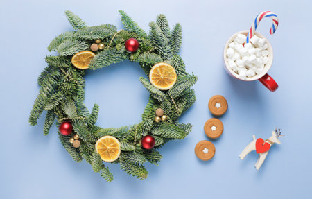 Christmas wreath made of fir branches, with small red and gold balls, slices of dried orange, shiny beads on a blue surface. A red cup of coffee with marshmallows, a candy cane and a deer figurine.の写真素材